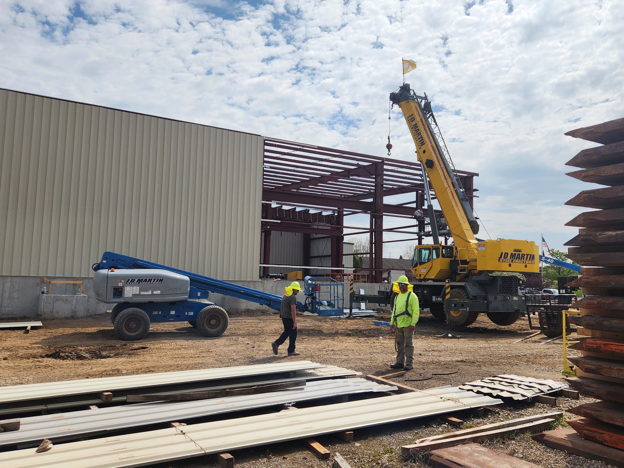 Construction workers operate cranes and lift equipment at a building site under a cloudy sky.