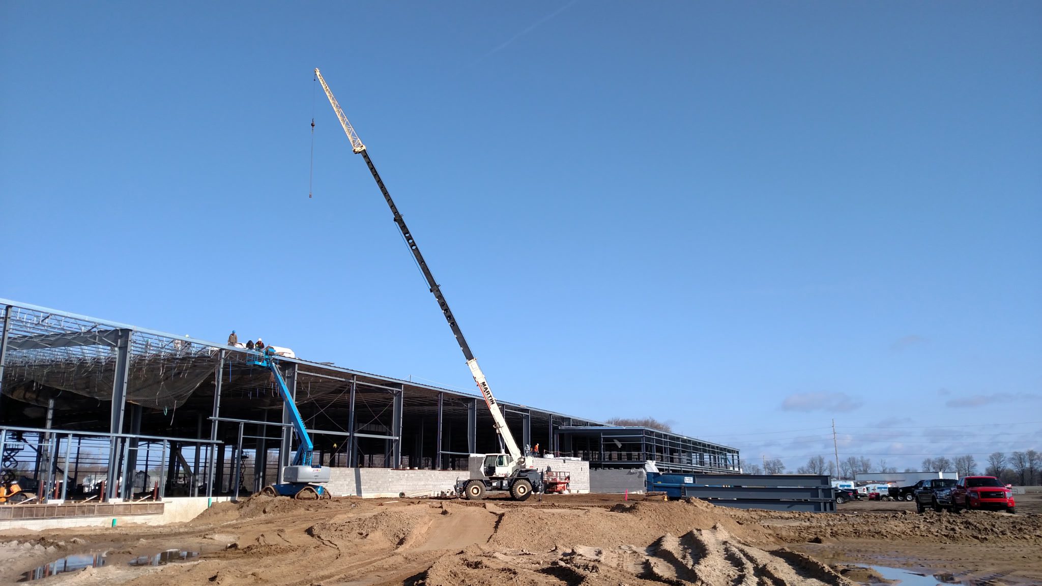 A crane lifts a large metal beam at a construction site under clear blue skies.