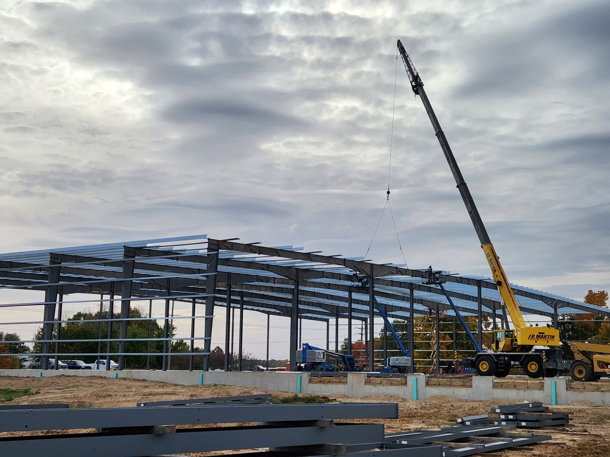 Steel framework being erected at a construction site with a crane under cloudy sky.