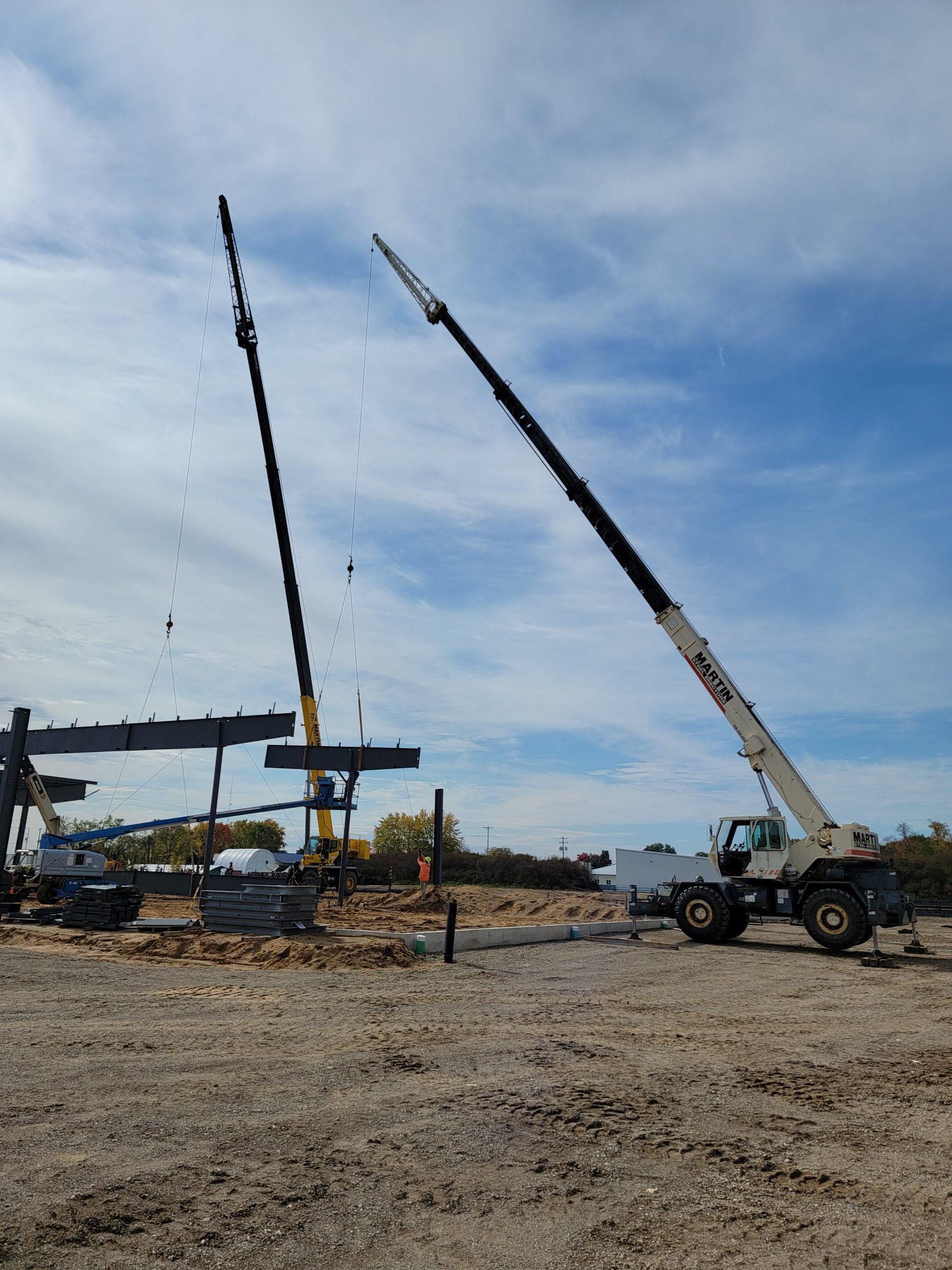 Two cranes lifting a large concrete beam at a construction site under a partly cloudy sky.